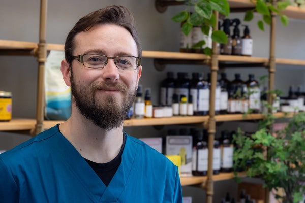 Smiling Quinn's Apothecary Pharmacy male staff member in front of wall of wellness products