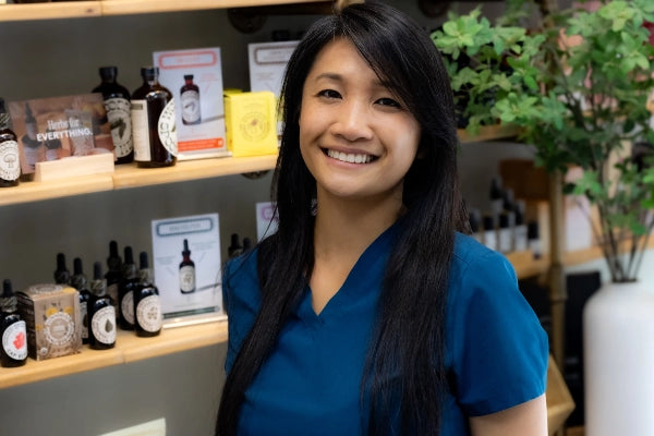 Smiling female pharmacist staff member at Quinn's Apothecary Pharmacy in front of wall of wellness products