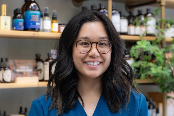 Smiling female pharmacist staff member at Quinn's Apothecary Pharmacy in front of wall of wellness products